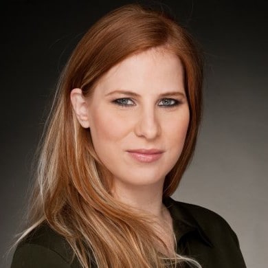 אדם עם שיער ערמוני ארוך וחלק לובש חלק עליון כהה, מתחזה על רקע אפור.
