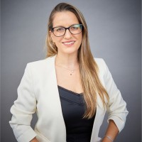 אדם עם שיער ארוך ומשקפיים, לבוש בלייזר לבן מעל חלק עליון שחור, מחייך על רקע אפור.